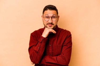 Latin kökenli genç adam bej arka planda izole edilmiş mutlu ve kendinden emin gülümsüyor, elleriyle çenesine dokunuyor..