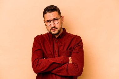 Latin kökenli genç adam bej arka planda izole edilmiş. Mutsuz, iğneleyici bir ifadeyle kameraya bakıyor..