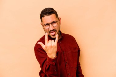 Latin kökenli genç adam bej arka planda izole edilmiş. Parmaklarıyla kaya hareketi gösteriyor.