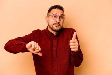 Latin kökenli genç bir adam bej arka planda izole edilmiş. Baş parmağını kaldırıyor ve baş parmağıyla işaret ediyor. Zor seçim kavramı.