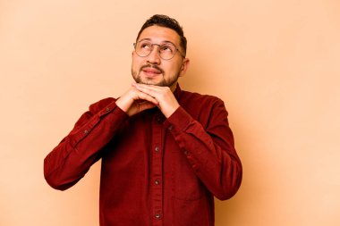 Bej arka planda izole edilmiş genç İspanyol adam ellerini çenesinin altında tutuyor ve mutlu bir şekilde yan tarafa bakıyor..