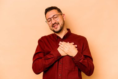 Latin kökenli genç bir adam bej arka planda izole edilmiş. Gülüyor, elini kalbe koyuyor, mutluluk kavramını koruyor..