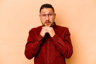 Latin kökenli genç adam bej arka planda izole edilmiş şans için dua ediyor, şaşırmış ve ağzını açıyor..