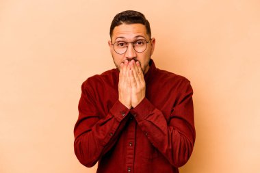 Latin kökenli genç bir adam, bej arka planda izole edilmiş şok olmuş, ağzı ellerle kaplanmış, yeni bir şey keşfetmeye hevesli..