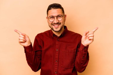 Latin kökenli genç adam bej arka planda izole edilmiş farklı fotokopi alanlarını gösteriyor, birini seçiyor, parmağıyla gösteriyor..