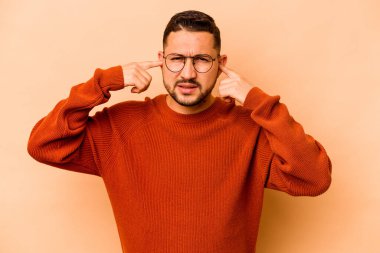 Latin kökenli genç adam bej arka planda izole edilmiş. Kulakları parmaklarıyla kapatıyor. Yüksek sesli bir ortam yüzünden stresli ve çaresiz..