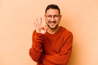 Latin kökenli genç bir adam bej arka planda izole edilmiş bir şekilde göz açıp kapayana kadar iyi bir el hareketi yapıyor..