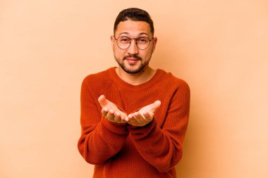 Latin kökenli genç adam bej arka planda izole edilmiş. Elinde avuç içi olan bir şey var ve kameraya sunuyor..