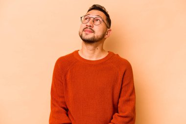 Latin kökenli genç adam bej arka planda izole edilmiş rahat ve mutlu bir şekilde gülüyor, boynunda dişlerini gösteriyor..