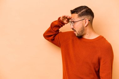 Latin kökenli genç adam bej arka planda izole edilmiş, uzaklara bakıyor ve elini alnına koyuyor..