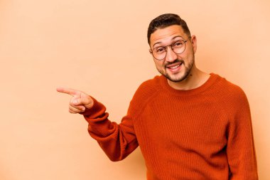 Latin kökenli genç adam, bej arka planda izole edilmiş. Gülümsüyor, işaret parmağıyla işaret ediyor..