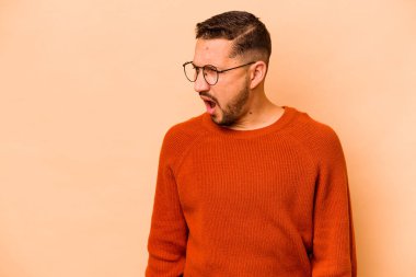 Latin kökenli genç adam bej arka planda izole edilmiş çok kızgın, öfkeli, hayal kırıklığına uğramış bağırıyor..