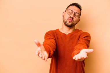 Latin kökenli genç adam, bej arka planda izole edilmiş dudakları kıvrılmış ve avuç içleriyle hava öpücüğü yolluyor..