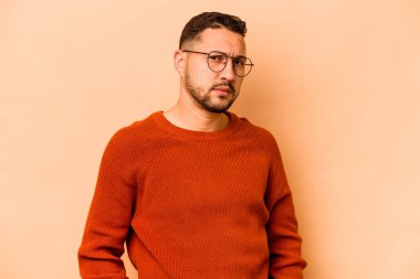 Latin kökenli genç adam bej arka planda izole edilmiş hoşnutsuzca kaşlarını çatmış, kollarını kavuşturmuş..