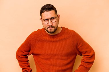 Latin kökenli genç adam bej arka planda izole edilmiş kafası karışmış, şüpheli ve güvensiz hissediyor..