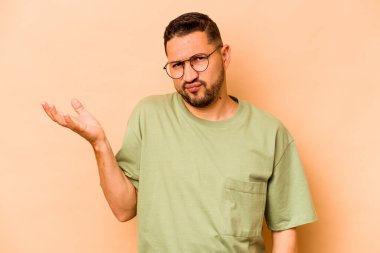 Latin kökenli genç bir adam, bej arka planda izole edilmiş sorgulama jestinde şüphe ediyor ve omuz silkiyor..