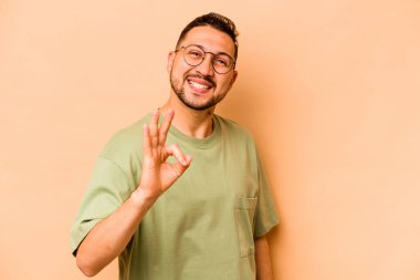 Latin kökenli genç adam bej arka planda izole edilmiş neşeli ve kendinden emin, iyi bir jest yapıyor..