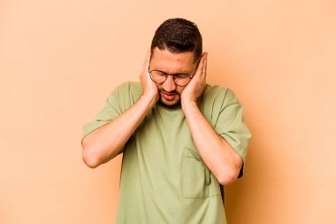 Latin kökenli genç adam bej arka planda izole edilmiş kulakları elleriyle kapatıyor..