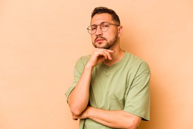 Latin kökenli genç bir adam, bej arka planda izole edilmiş şüpheli, belirsiz, seni muayene ediyor..