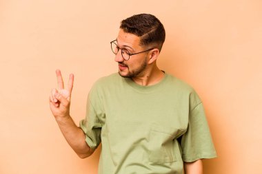 Genç İspanyol adam bej arka planda izole edilmiş neşeli ve kaygısız parmakları olan bir barış sembolü gösteriyor..