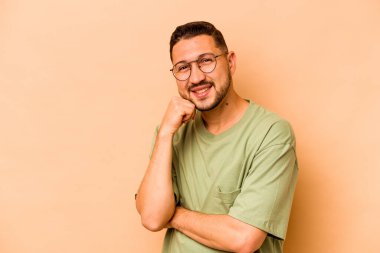 Latin kökenli genç adam bej arka planda izole edilmiş mutlu ve kendinden emin gülümsüyor, elleriyle çenesine dokunuyor..