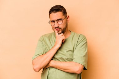 Latin kökenli genç adam bej arka planda izole edilmiş. Mutsuz, iğneleyici bir ifadeyle kameraya bakıyor..