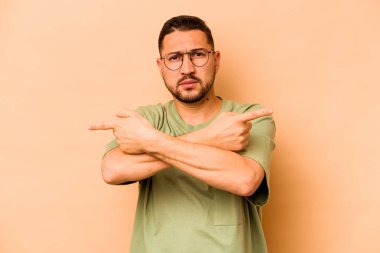 Latin kökenli genç adam bej arka planda iki seçenek arasında seçim yapmaya çalışıyor..