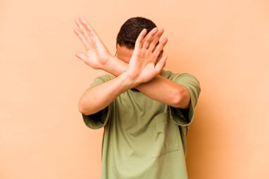 Latin kökenli genç adam bej arka planda izole edilmiş iki kolu çapraz tutuyor, inkar kavramı.