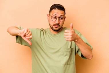 Latin kökenli genç bir adam bej arka planda izole edilmiş. Baş parmağını kaldırıyor ve baş parmağıyla işaret ediyor. Zor seçim kavramı.