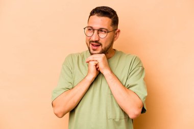 Bej arka planda izole edilmiş genç İspanyol adam ellerini çenesinin altında tutuyor ve mutlu bir şekilde yan tarafa bakıyor..