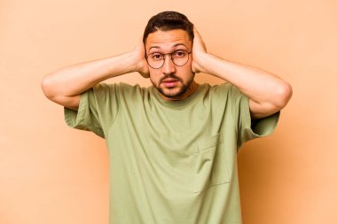 Latin kökenli genç adam bej arka planda izole edilmiş kulakları kulaklarıyla kapatıyor çok fazla ses duymamaya çalışıyor..