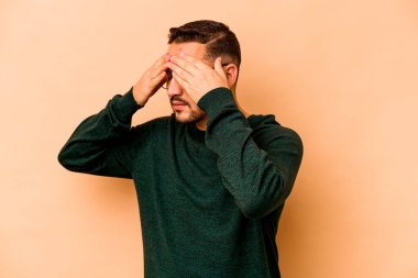 Latin kökenli genç bir adam bej arka planda izole edilmiş, elleriyle gözlerini kapatmaktan korkuyor..