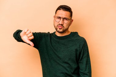 Latin kökenli genç adam bej arka planda izole edilmiş baş parmağını aşağı gösteren, hayal kırıklığı konsepti.