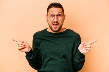 Latin kökenli genç adam bej arka planda izole edilmiş farklı fotokopi alanlarını gösteriyor, birini seçiyor, parmağıyla gösteriyor..