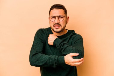 Genç İspanyol adam bej arka planda izole edilmiş. Dirsek masajı yapıyor. Kötü bir hareketin ardından acı çekiyor..