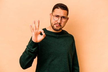 Latin kökenli genç bir adam bej arka planda izole edilmiş bir şekilde göz açıp kapayana kadar iyi bir el hareketi yapıyor..