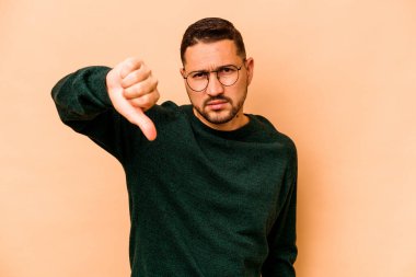 Latin kökenli genç adam bej arka planda izole edilmiş baş parmağını aşağı gösteriyor ve hoşlanmadığını ifade ediyor..