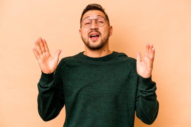 Latin kökenli genç adam bej arka planda izole edilmiş. Yüksek sesle gülüyor. Elini göğsünde tutuyor..