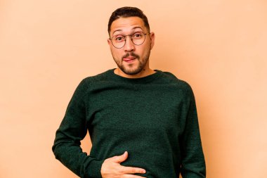 Latin kökenli genç adam bej arka planda izole edilmiş karnına dokunuyor, nazikçe gülümsüyor, yemek yiyor ve tatmin oluyor..