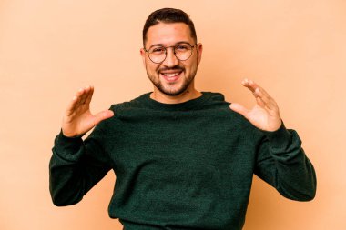 Latin kökenli genç adam bej arka planda izole edilmiş. Elinde avuç içi olan bir şey var ve kameraya sunuyor..