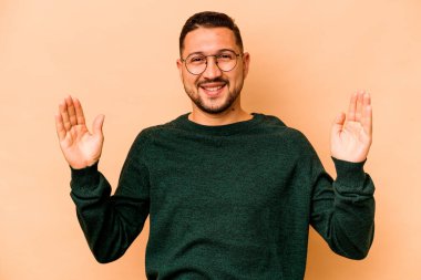 Latin kökenli genç adam bej arka planda izole edilmiş. Elinde küçük bir işaret parmağı olan, gülümseyen ve kendinden emin bir şey tutuyor..