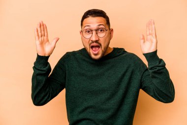 Latin kökenli genç bir adam bej arka planda izole edilmiş. Hoş bir sürpriz alıyor, heyecanlı ve el kaldırıyor..