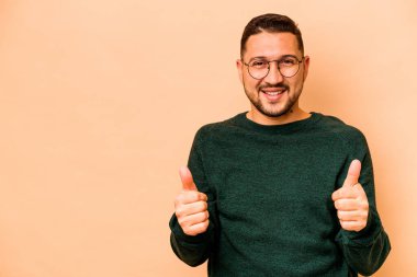 Genç İspanyol adam bej arka planda izole edilmiş gülümsüyor ve başparmağını kaldırıyor.