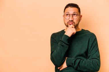 Latin kökenli genç adam bej arka planda izole edilmiş şüpheli ve şüpheci bir ifadeyle yana bakıyor..