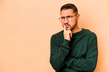 Latin kökenli genç adam bej arka planda izole edilmiş şüpheli ve şüpheci bir ifadeyle yana bakıyor..