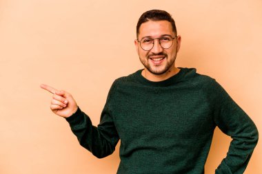 Latin kökenli genç adam, bej arka planda izole edilmiş. Gülümsüyor, işaret parmağıyla işaret ediyor..
