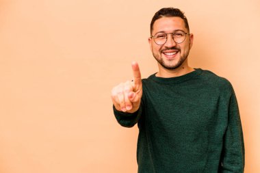 Latin kökenli genç bir adam bej arka planda izole edilmiş. Parmağıyla bir numarayı gösteriyor..
