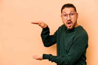 Latin kökenli genç adam bej arka planda izole edilmiş. Elleri arasında bir kopya alanı tutarken şok olmuş ve hayrete düşmüştü..