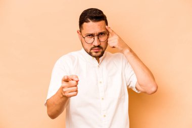 Latin kökenli genç bir adam bej arka planda izole edilmiş. Şakağı parmağıyla işaret ediyor, düşünüyor ve bir göreve odaklanıyor..