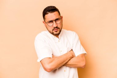 Latin kökenli genç adam bej arka planda izole edilmiş hoşnutsuzca kaşlarını çatmış, kollarını kavuşturmuş..